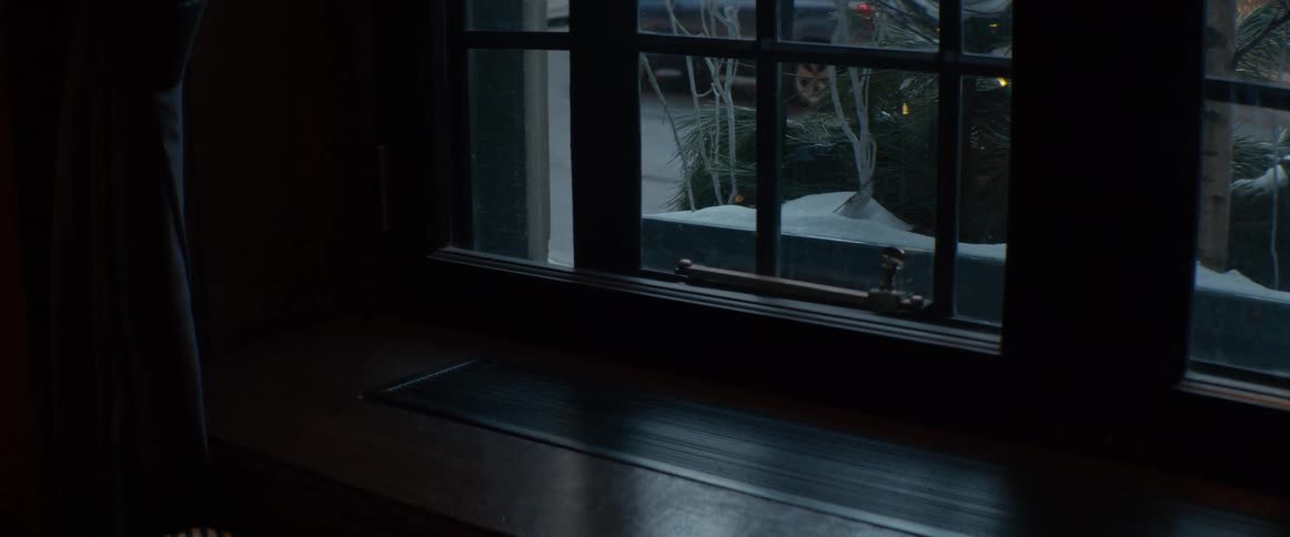 Looking out a dark wooden window from a cozy room onto a snowy street scene at dusk