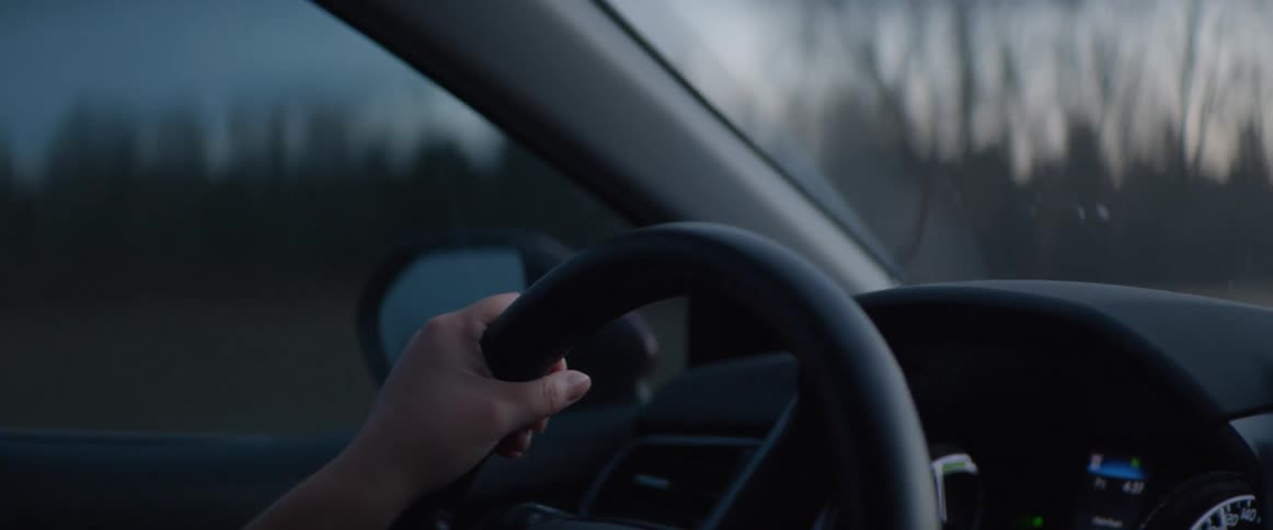 A person's hand firmly holds the steering wheel while driving a car on a highway during the evening commute