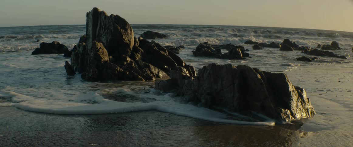 Slow motion shot of gentle ocean waves washing over dark, sunlit rock formations on a sandy beach at golden hour