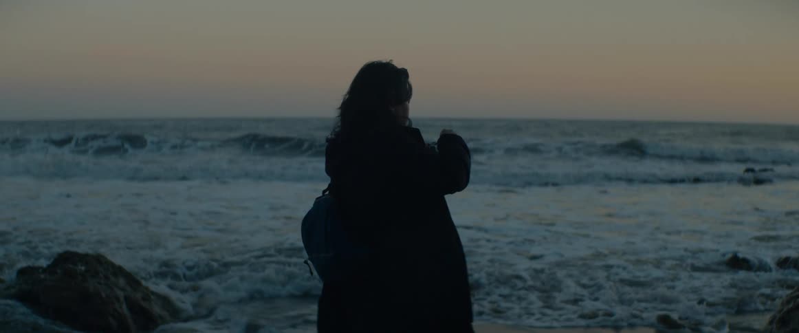 A silhouetted woman with a backpack takes photos of the rolling ocean waves during a peaceful sunrise