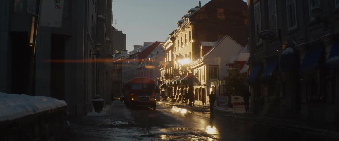 A golden hour sunbeam illuminates a snowy downtown city street with a parked school bus and people walking past shops