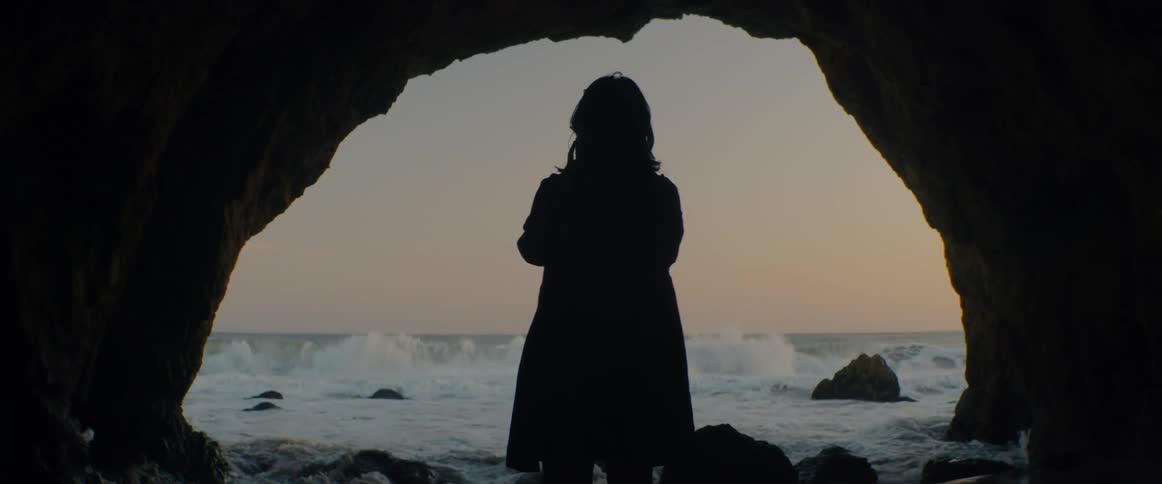 A silhouetted figure stands at the mouth of a sea cave, looking out at the ocean as waves roll in at sunset