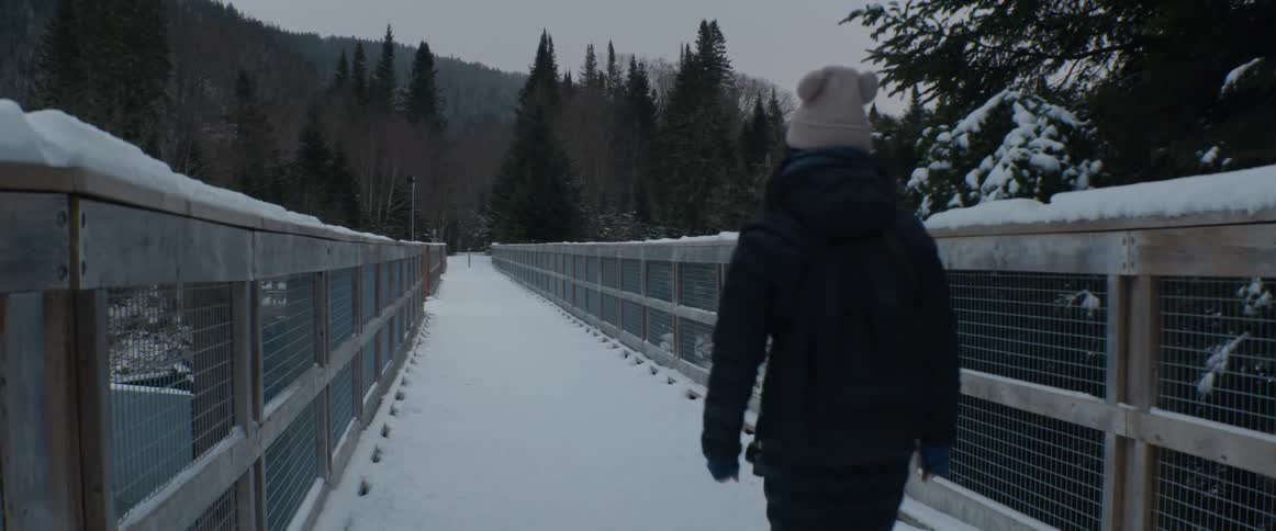 A woman wearing a winter coat and beanie walks away from the camera down a long, snow-covered bridge in a remote forest