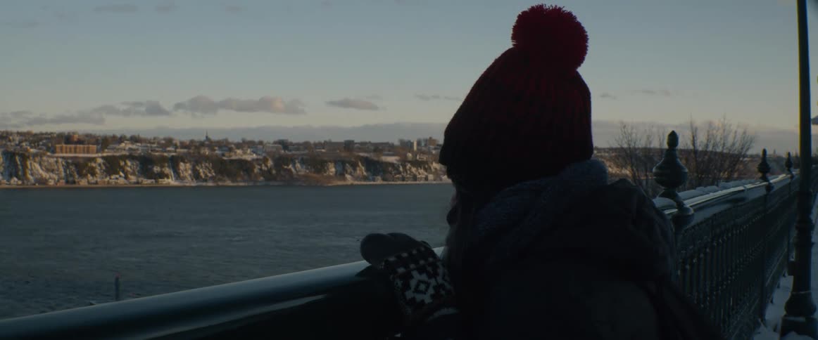 A solitary figure in a winter beanie stands at a railing, quietly observing a snowy city across a river during a peaceful sunset