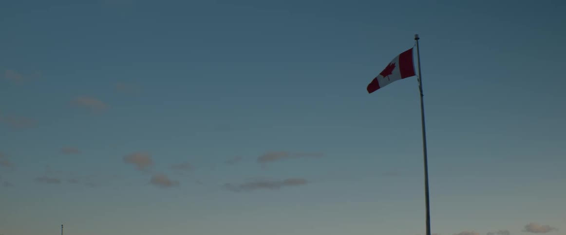 A Canadian flag waves gently on a flagpole against a clear blue sky at twilight