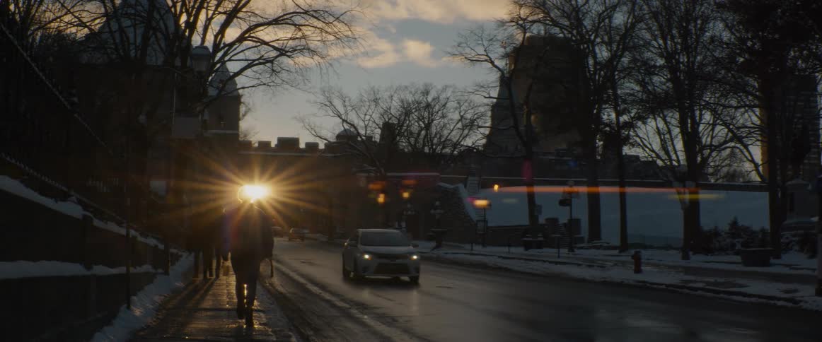 A person walks on a snowy sidewalk in a historic city as the setting sun creates a beautiful lens flare while a car drives by