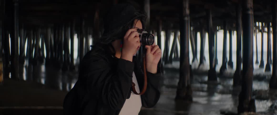 A woman with a vintage-style camera takes photos of the ocean while standing under a dark pier