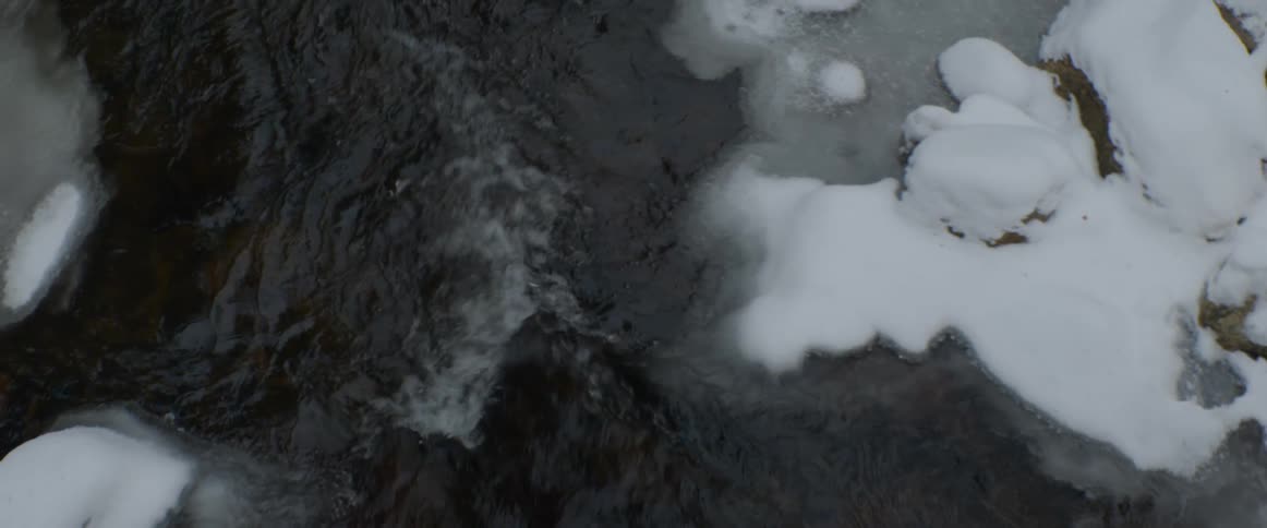 Dark water flows past snow and ice-covered rocks in a winter stream