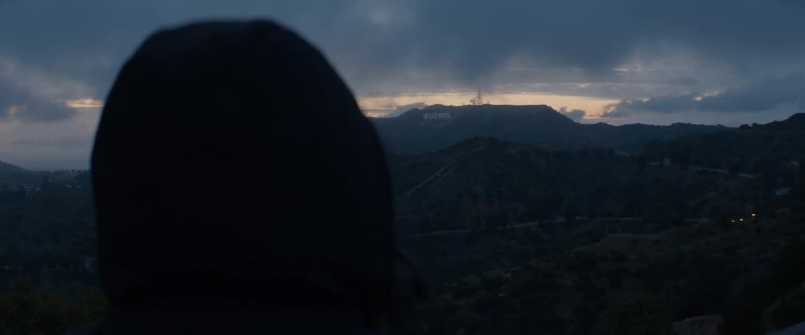 A person in a dark hoodie looks out at the iconic Hollywood sign from a viewpoint in the hills at dusk
