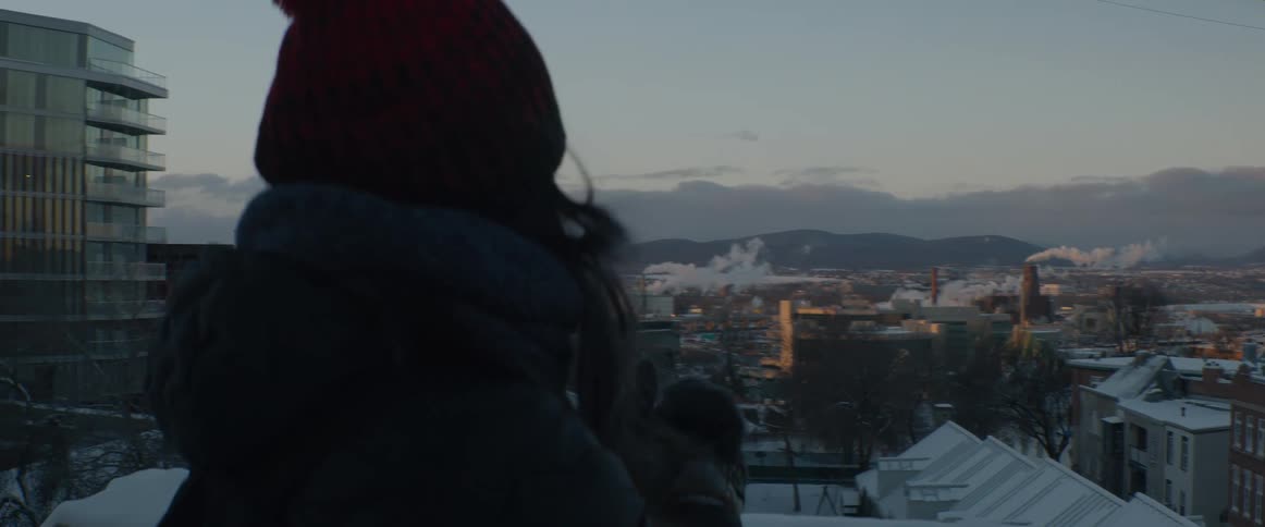 A woman in a winter coat and beanie looks out over a snowy city with mountains in the distance at dawn