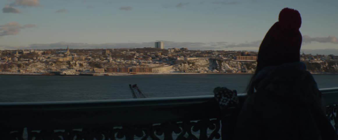 A person in a red beanie and winter clothes gazes out over a river at a snow-covered city during a beautiful sunrise