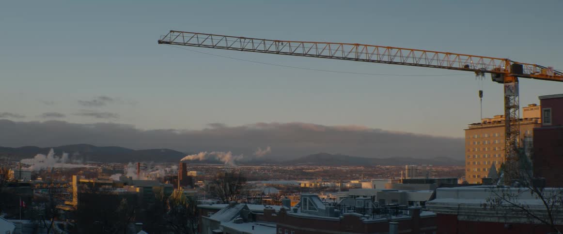 Sunrise over an industrial city skyline featuring a prominent construction crane and smoke billowing from chimneys