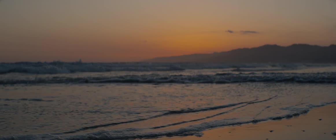 Slow motion footage shows waves gently lapping on a sandy beach, reflecting the warm glow of the setting sun