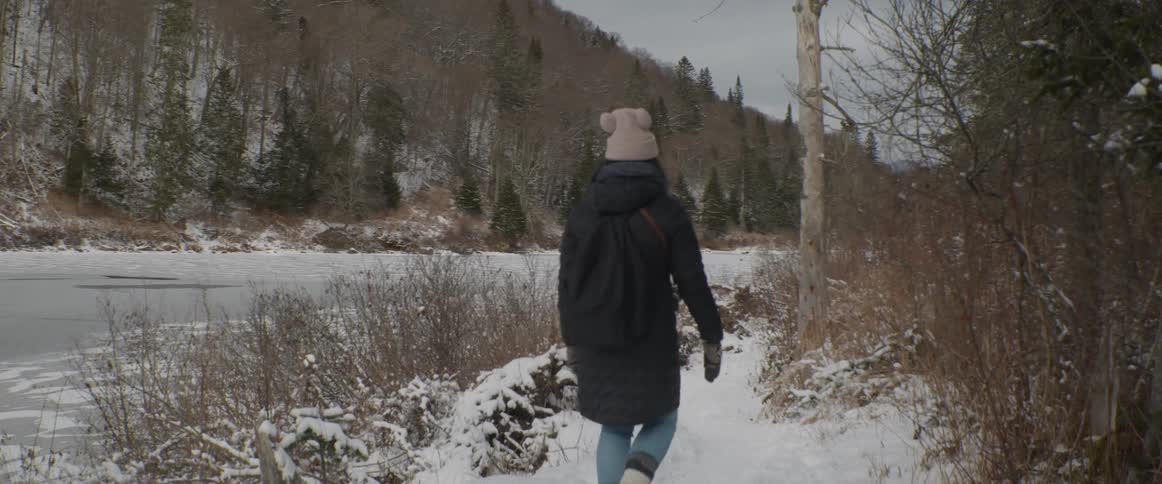 A woman wearing a cute pom-pom hat walks in slow motion along a snowy path next to a frozen river in a wintery forest