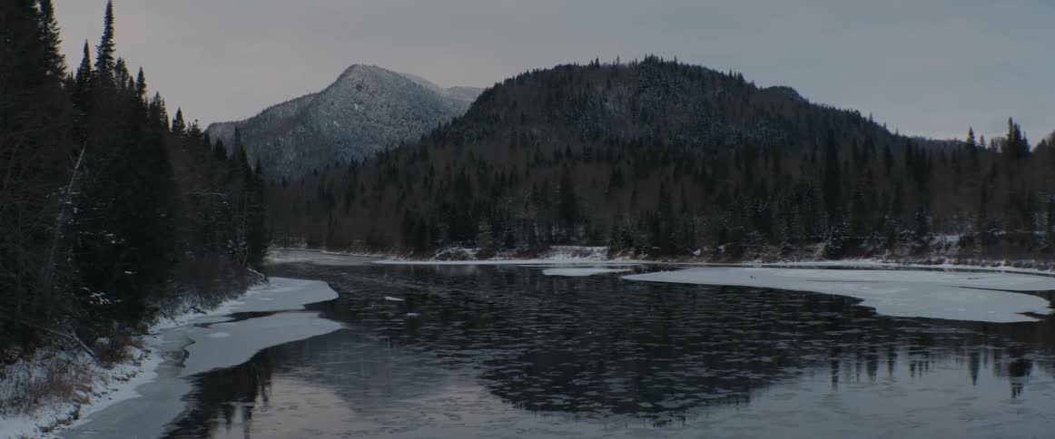 A partially frozen river reflects snow-dusted mountains and a dense evergreen forest on a calm, overcast winter day