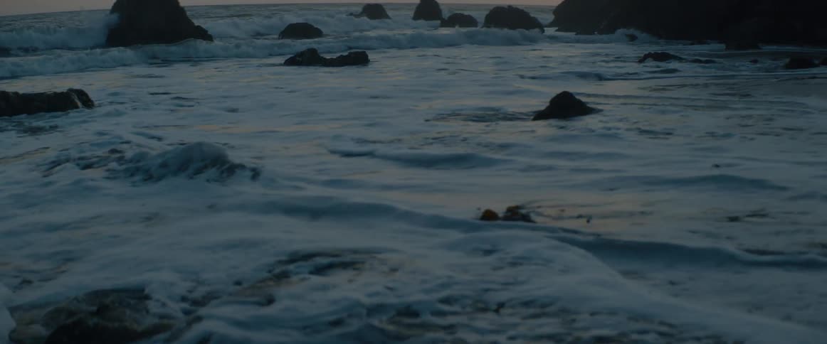 A slow-motion close-up captures foamy ocean waves receding from a rocky shoreline at dusk