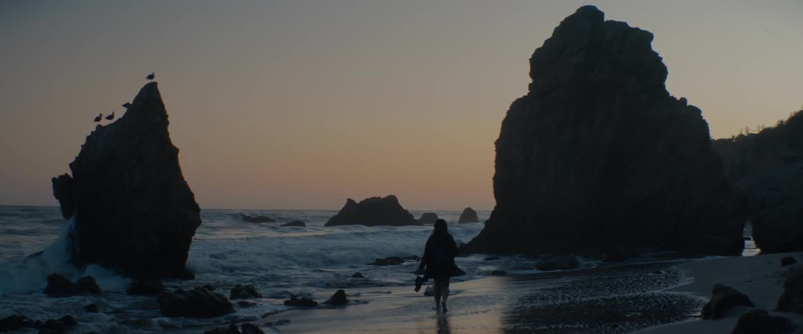 A solitary figure walks barefoot on a sandy beach between large rock formations during a peaceful sunset