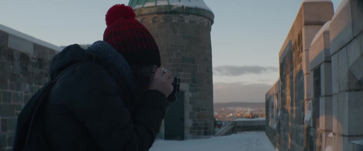 A young woman wearing a red beanie takes a photograph of the cityscape from a snowy historic fortress at sunset
