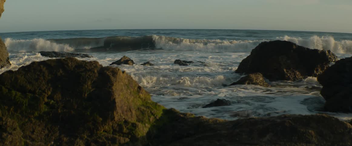 Serene slow-motion view of ocean waves rolling and crashing against mossy rocks along the shoreline at sunset
