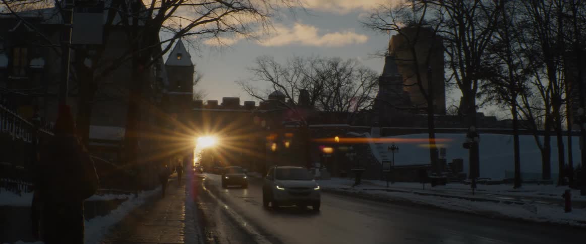 The setting sun shines through a historic city gate, creating a brilliant lens flare as cars drive on a snowy winter road