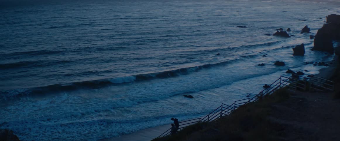 A silhouetted couple stands on beach stairs watching the ocean waves roll in at twilight