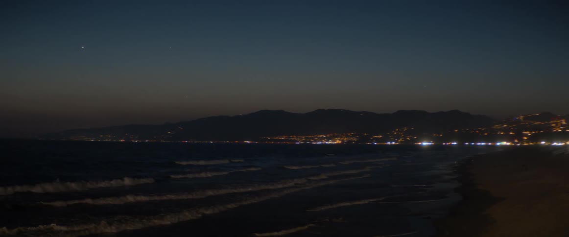 Ocean waves wash ashore at dusk with the sparkling lights of a coastal city and mountains in the distance