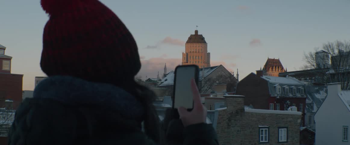 A tourist in a red beanie uses a smartphone to capture the snowy Quebec City skyline at sunrise