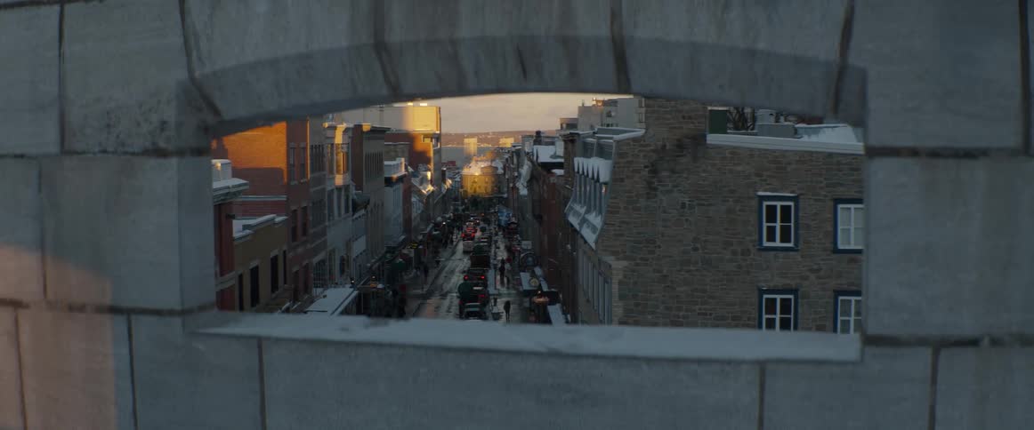 A picturesque view through a stone arch reveals a busy, snow-covered city street with cars and pedestrians during a golden hour sunset