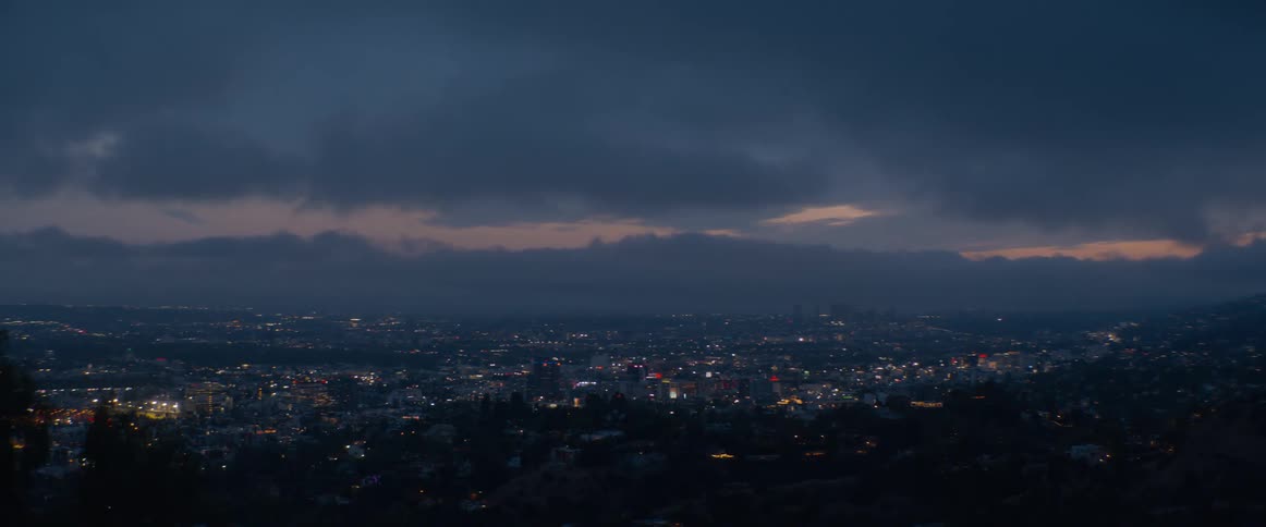 The sprawling city lights of Los Angeles glitter under a dark and cloudy sky at dusk