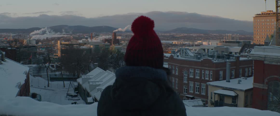 A person in a red knit hat enjoys the view of a snowy city landscape with mountains in the distance at sunrise