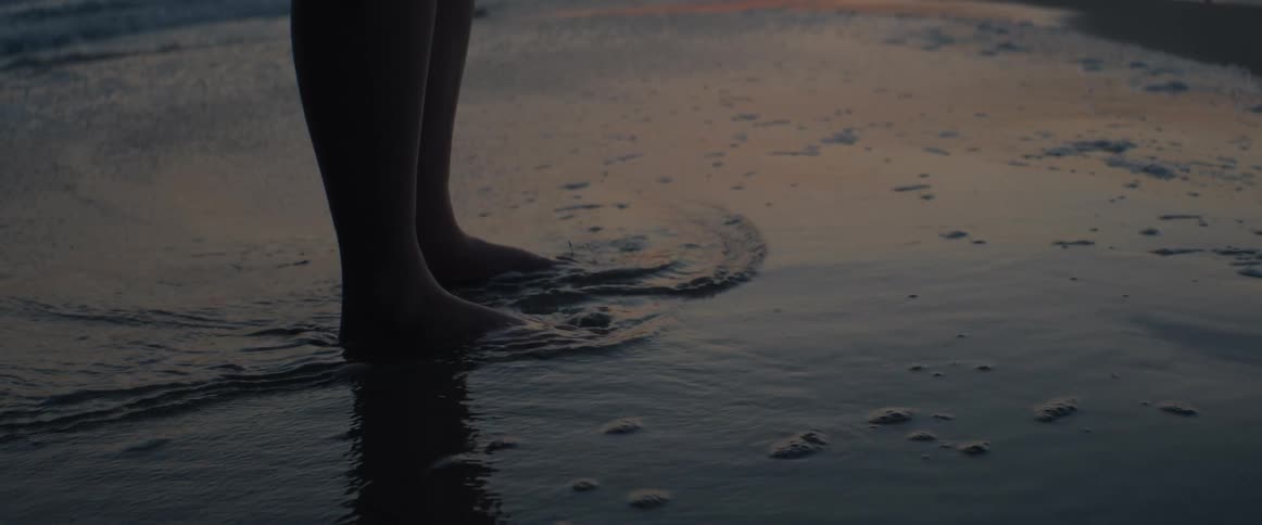 The silhouette of a person's bare feet are bathed by gentle ocean waves at sunset in slow motion