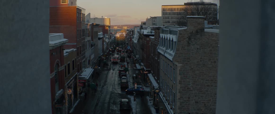 A high-angle view reveals a bustling city street lined with historic, snow-covered buildings as traffic flows during a winter sunset