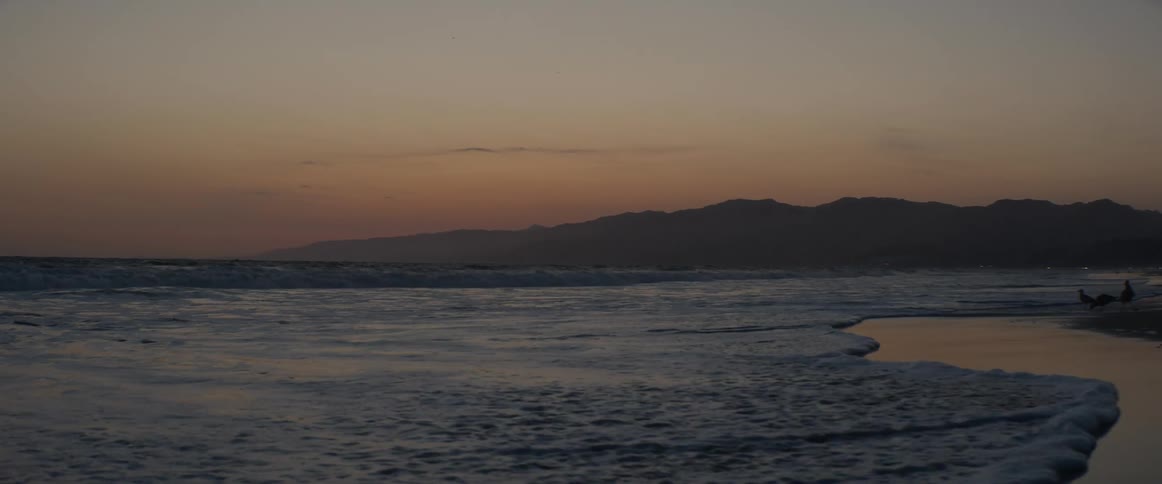 Calm ocean waves gently roll onto a sandy shore in slow motion during a picturesque sunset with mountains on the horizon