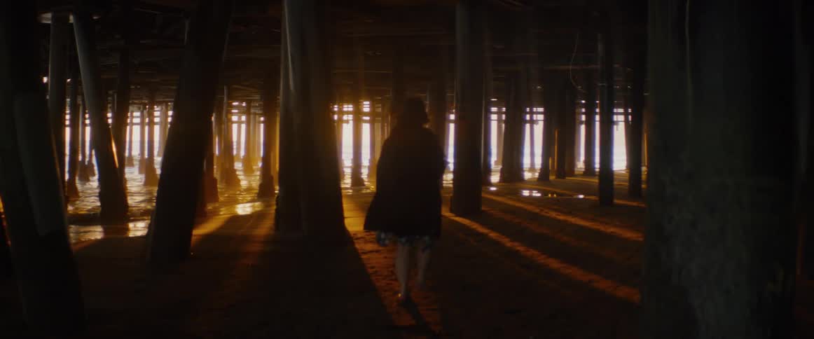 A silhouetted woman walks slowly and barefoot on the sand under a pier as the golden sun sets over the ocean