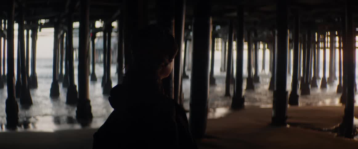 A silhouetted figure stands under a dark pier and watches the gentle ocean waves roll in