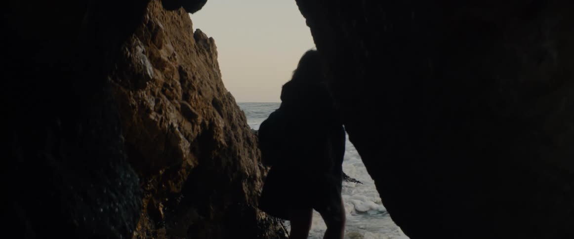 From within a dark sea cave, a silhouetted woman walks out towards the crashing ocean waves