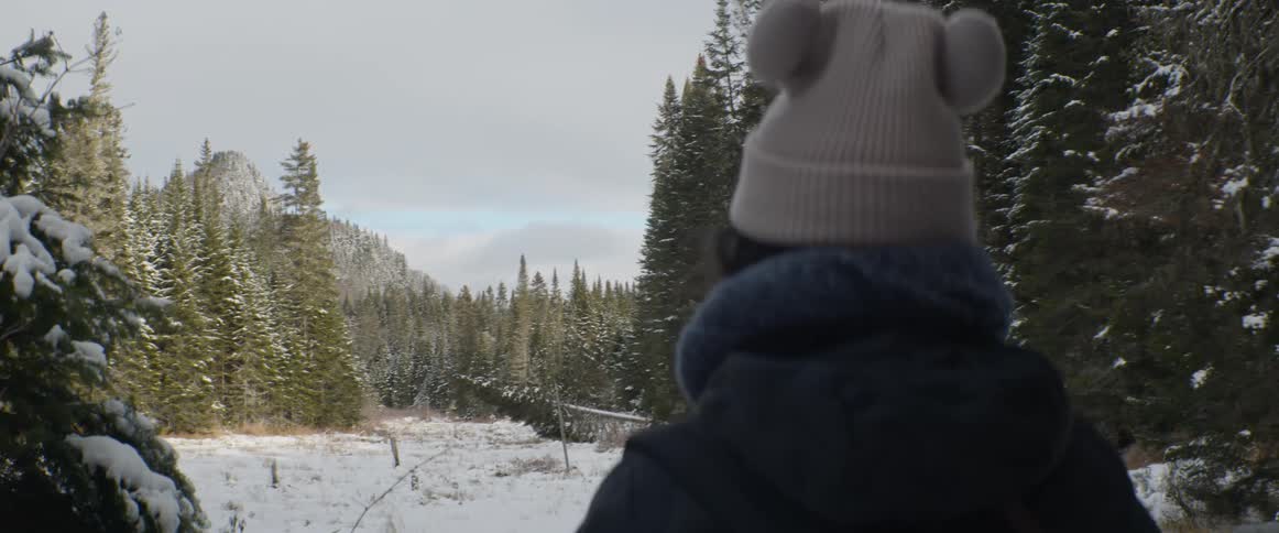 Over the shoulder view of a person in a pom-pom beanie looking out at a scenic snowy mountain forest