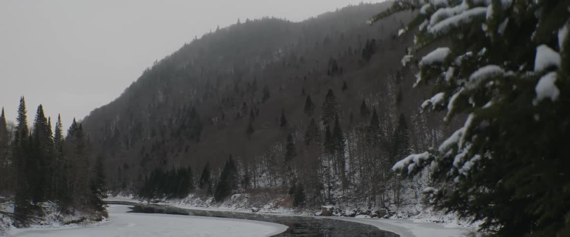 A tranquil winter landscape shows a partially frozen river meandering through a snowy forest at the foot of a mountain