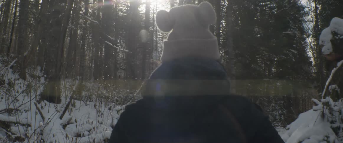 A person wearing a playful pom-pom beanie walks through a snowy forest wonderland as sunlit snowflakes gently fall