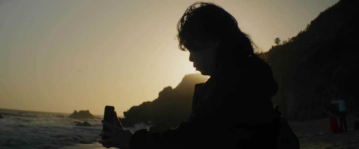 A silhouetted young person with long hair captures the beautiful golden hour sunset over the ocean on their smartphone