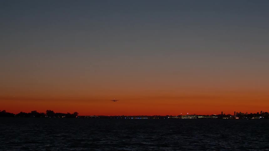 An airplane with its lights on descends for landing at a distant airport over the water during a beautiful orange sunset