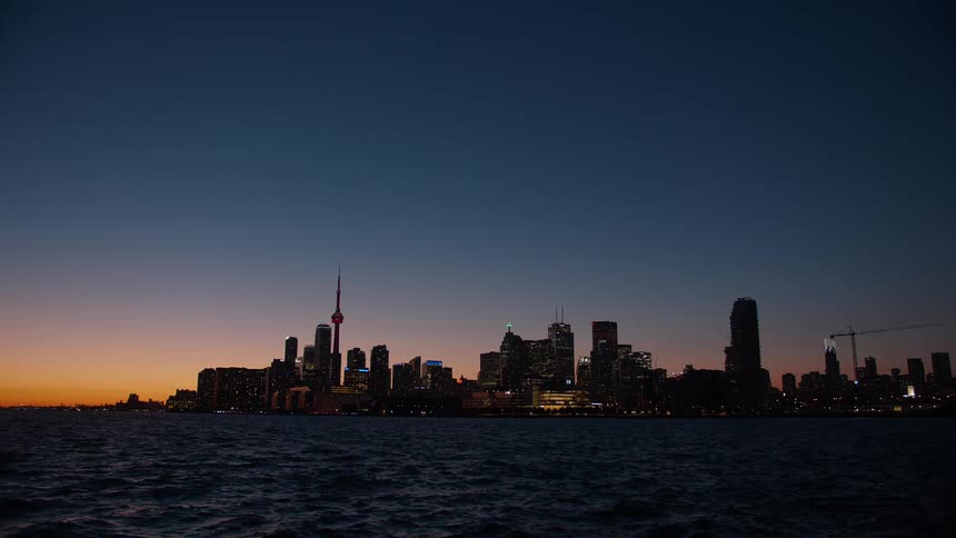 Calm waters ripple in front of the iconic Toronto skyline as city lights twinkle under a beautiful sunset