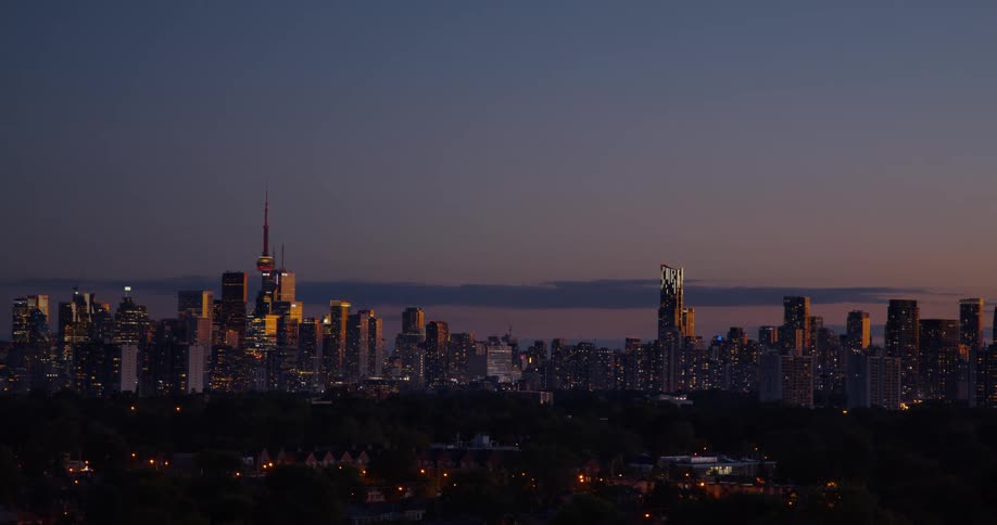 The glowing skyline of downtown Toronto is seen from a distance at twilight as night falls
