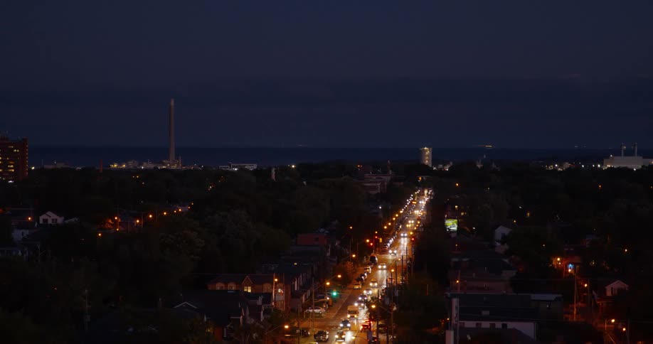 A long, illuminated city street with heavy traffic stretches towards the distant waterfront at dusk