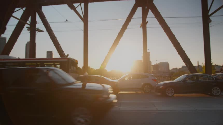 Golden hour sunset reflects off cars and a city bus moving slowly through rush hour traffic on a steel truss bridge