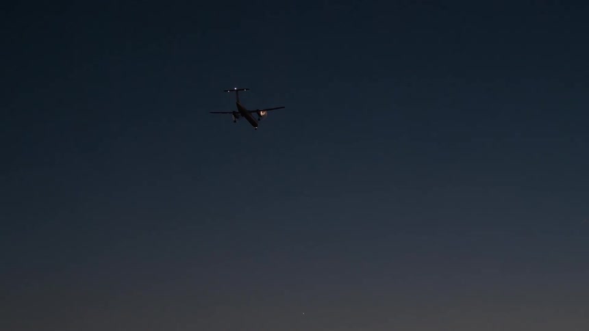 A close-up shot of an airplane flying at night zooms out to reveal a beautiful sunset over the water as it approaches for landing