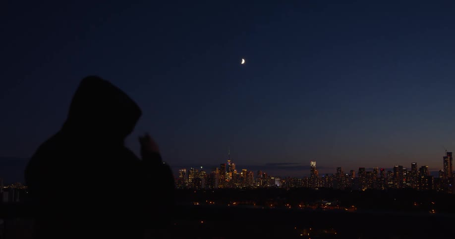 A silhouetted person contemplates the illuminated city skyline and crescent moon at twilight before pulling up their hood and leaving