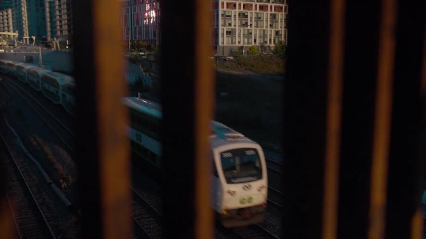 Viewed through silhouetted bars, a passenger train travels along urban tracks with city buildings in the background at sunset