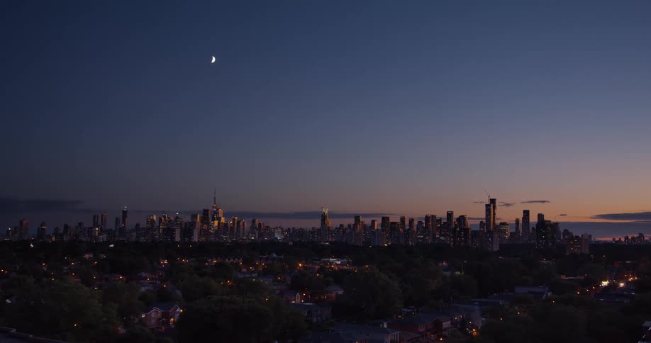 A crescent moon hangs in the dusky sky over an illuminated city skyline and a quiet residential neighborhood
