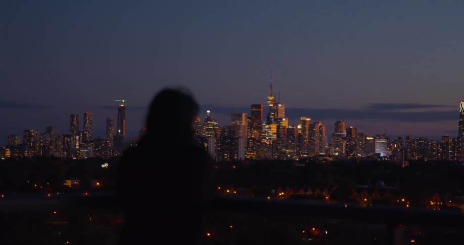 A lone figure stands silhouetted against the breathtaking Toronto skyline, illuminated by city lights at dusk